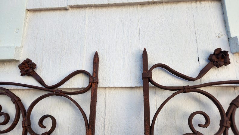Pu&ograve; includere: Primo piano di una sezione di un cancello in ferro battuto con finitura marrone ruggine. Il cancello presenta volute decorative, pinnacoli appuntiti e accenti floreali. Lo sfondo &egrave; una parete di legno bianco.