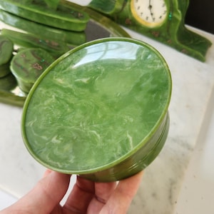 May include: A green oval trinket box with a marbled pattern, held in a hand. Other green decorative items are in the background, including a clock and a tray with matching boxes. The items have a vintage aesthetic.