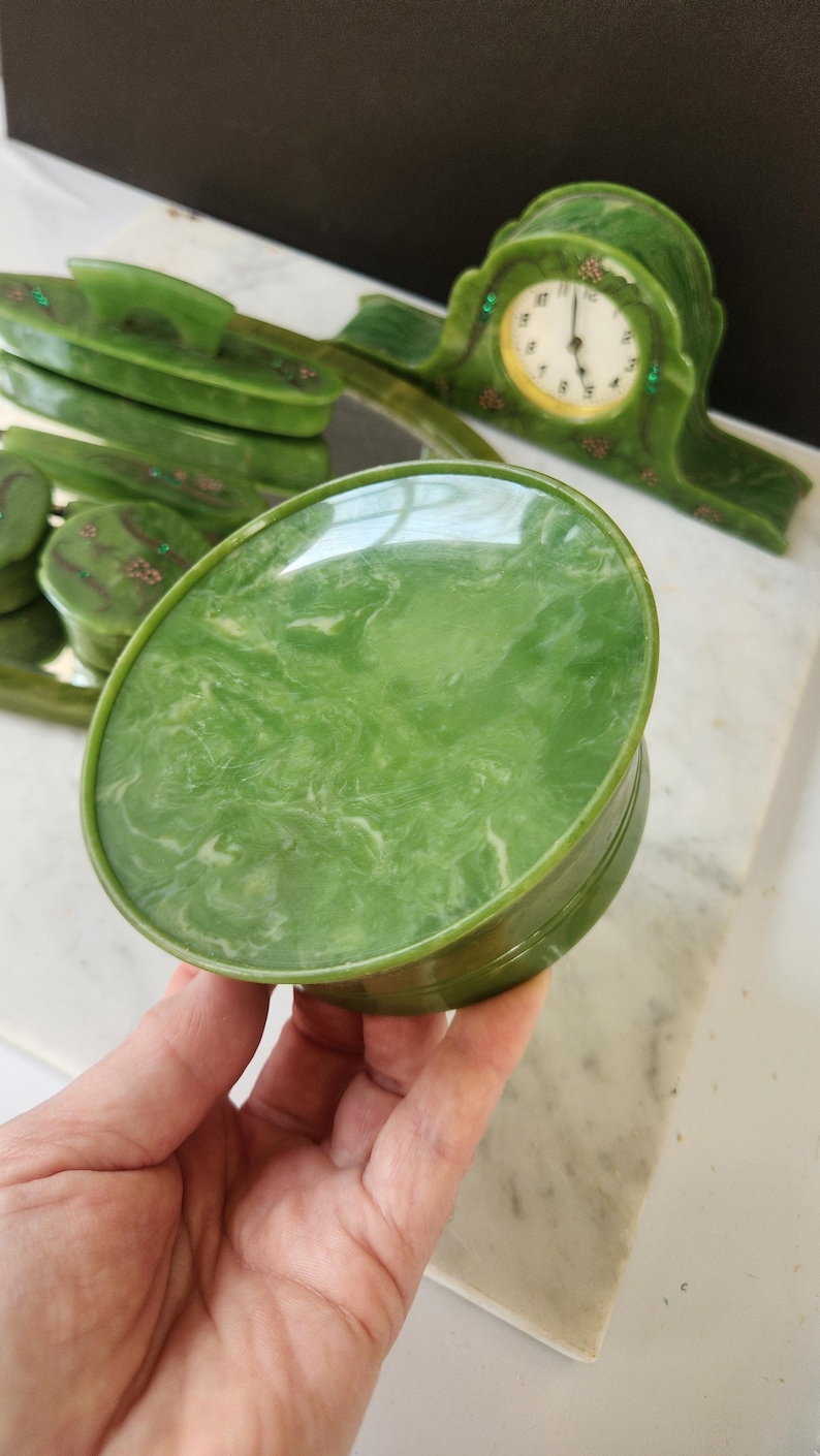 May include: A green oval trinket box with a marbled pattern, held in a hand. Other green decorative items are in the background, including a clock and a tray with matching boxes. The items have a vintage aesthetic.