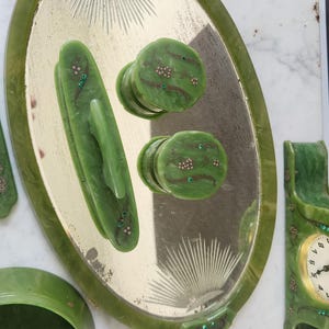 May include: A collection of green vintage vanity accessories. Includes an oval tray with a mirrored surface, two drawer pulls, a handle, a small bowl, and a clock. The pieces are decorated with small floral accents and green gemstones.