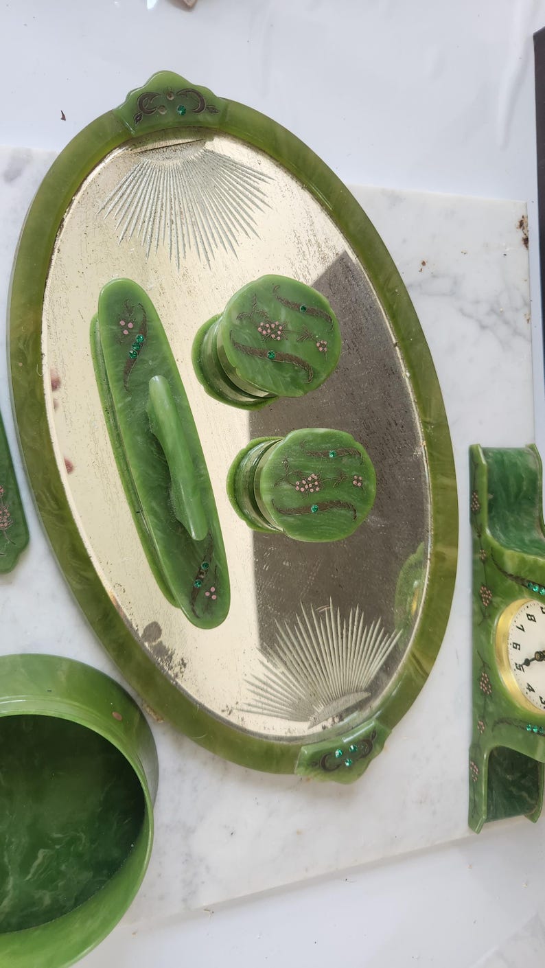May include: A collection of green vintage vanity accessories. Includes an oval tray with a mirrored surface, two drawer pulls, a handle, a small bowl, and a clock. The pieces are decorated with small floral accents and green gemstones.