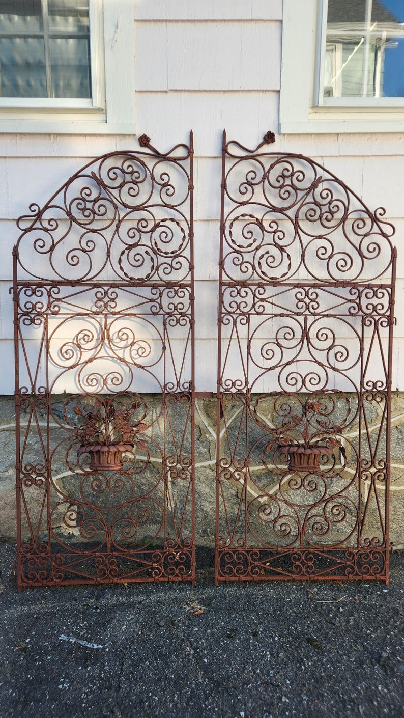 Pu&ograve; includere: Due cancelli ornamentali in ferro battuto color ruggine. Ogni cancello presenta un'intricata lavorazione a volute, accenti floreali e una fioriera decorativa. I cancelli sono progettati con un telaio rettangolare e una parte superiore ad arco, adatti per un giardino o un ingresso.