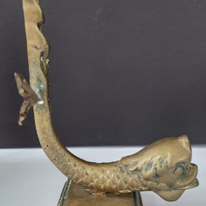 May include: A vintage brass dolphin-shaped object with a rectangular base. The dolphin curves upwards, with a detailed head and tail. The brass has a weathered appearance, with some green oxidation. The object is likely a decorative piece.