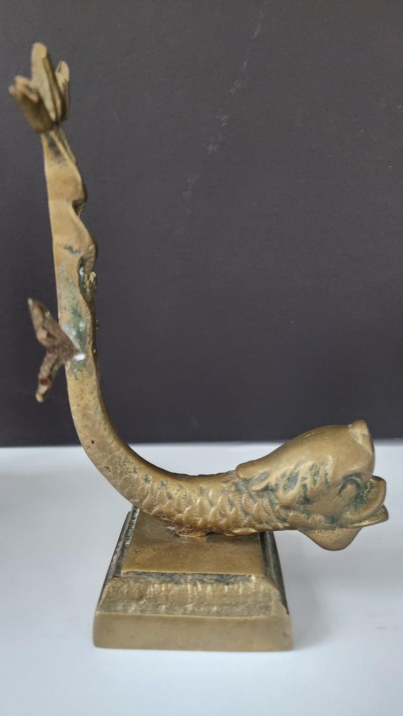 May include: A vintage brass dolphin-shaped object with a rectangular base. The dolphin curves upwards, with a detailed head and tail. The brass has a weathered appearance, with some green oxidation. The object is likely a decorative piece.