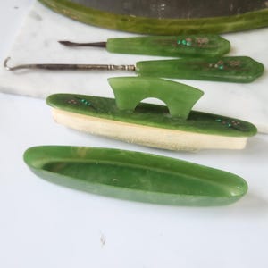 May include: A vintage green vanity set comprising a tray, jars, and grooming tools. The set has a green colour scheme with decorative accents. The items are arranged on a white surface, showcasing the elegant design of the vanity set.