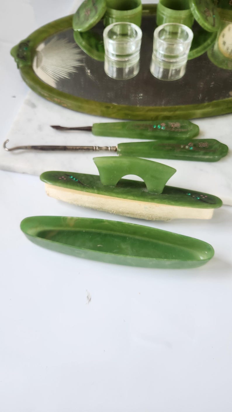 May include: A vintage green vanity set comprising a tray, jars, and grooming tools. The set has a green colour scheme with decorative accents. The items are arranged on a white surface, showcasing the elegant design of the vanity set.
