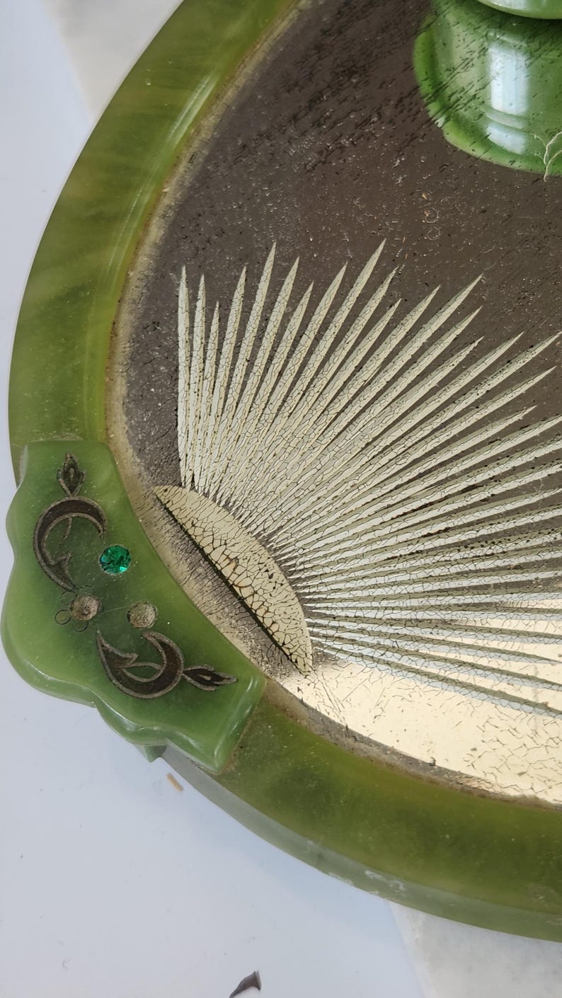 May include: A vintage green jade-coloured tray with a decorative design. The tray features a sunburst pattern in the centre with a gold-coloured finish. The rim is a darker green, and there is a decorative corner with a green gem.