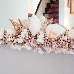 May include: A decorative mirror frame adorned with a variety of seashells, coral, and small, round, beige-colored shells. The seashells are in shades of white, beige, and brown, creating a coastal theme. The mirror reflects a room interior.