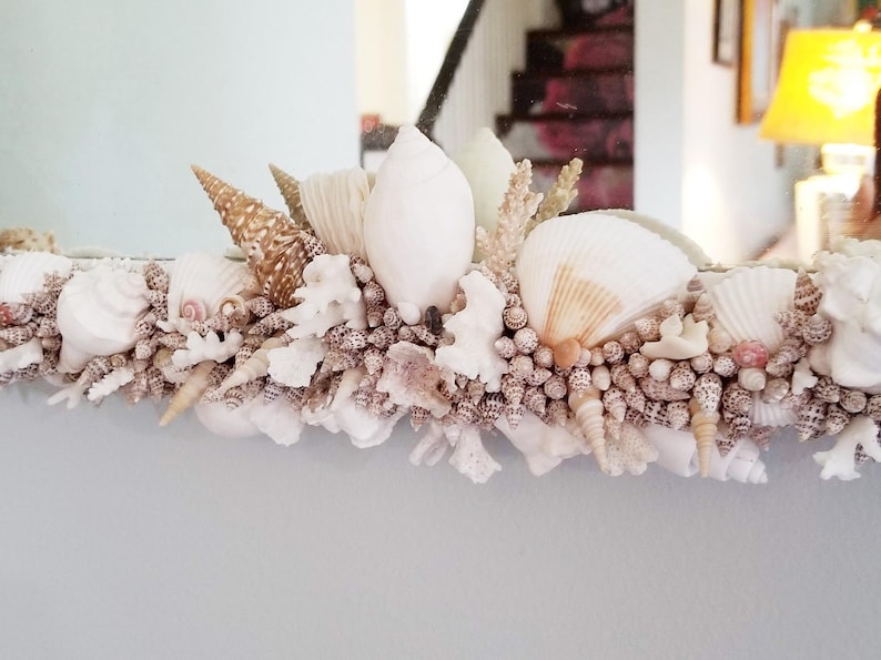 May include: A decorative mirror frame adorned with a variety of seashells, coral, and small, round, beige-colored shells. The seashells are in shades of white, beige, and brown, creating a coastal theme. The mirror reflects a room interior.
