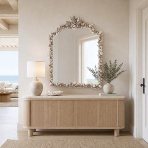 May include: A large, arched mirror framed with seashells hangs above a light wood cabinet. A lamp with a beige shade and a white vase with greenery sit on the cabinet. The room has a neutral color palette with a coastal theme.