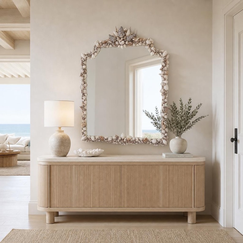 May include: A large, arched mirror framed with seashells hangs above a light wood cabinet. A lamp with a beige shade and a white vase with greenery sit on the cabinet. The room has a neutral color palette with a coastal theme.