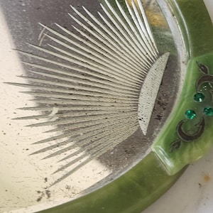 May include: A vintage vanity tray in green and silver, featuring a sunburst design. The tray has a rounded shape with a green border and decorative details. The silver surface shows signs of wear, enhancing its antique appeal.