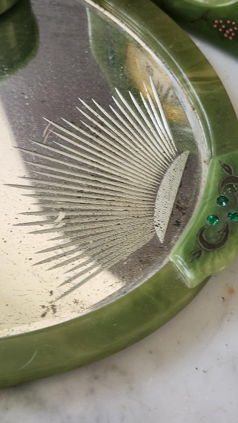 May include: A vintage vanity tray in green and silver, featuring a sunburst design. The tray has a rounded shape with a green border and decorative details. The silver surface shows signs of wear, enhancing its antique appeal.