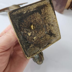May include: A close-up view of a vintage brass bird figurine base. The square base shows signs of age with dark patina and a central screw. The bird's feet are visible, adding to the antique aesthetic. The background is blurred.