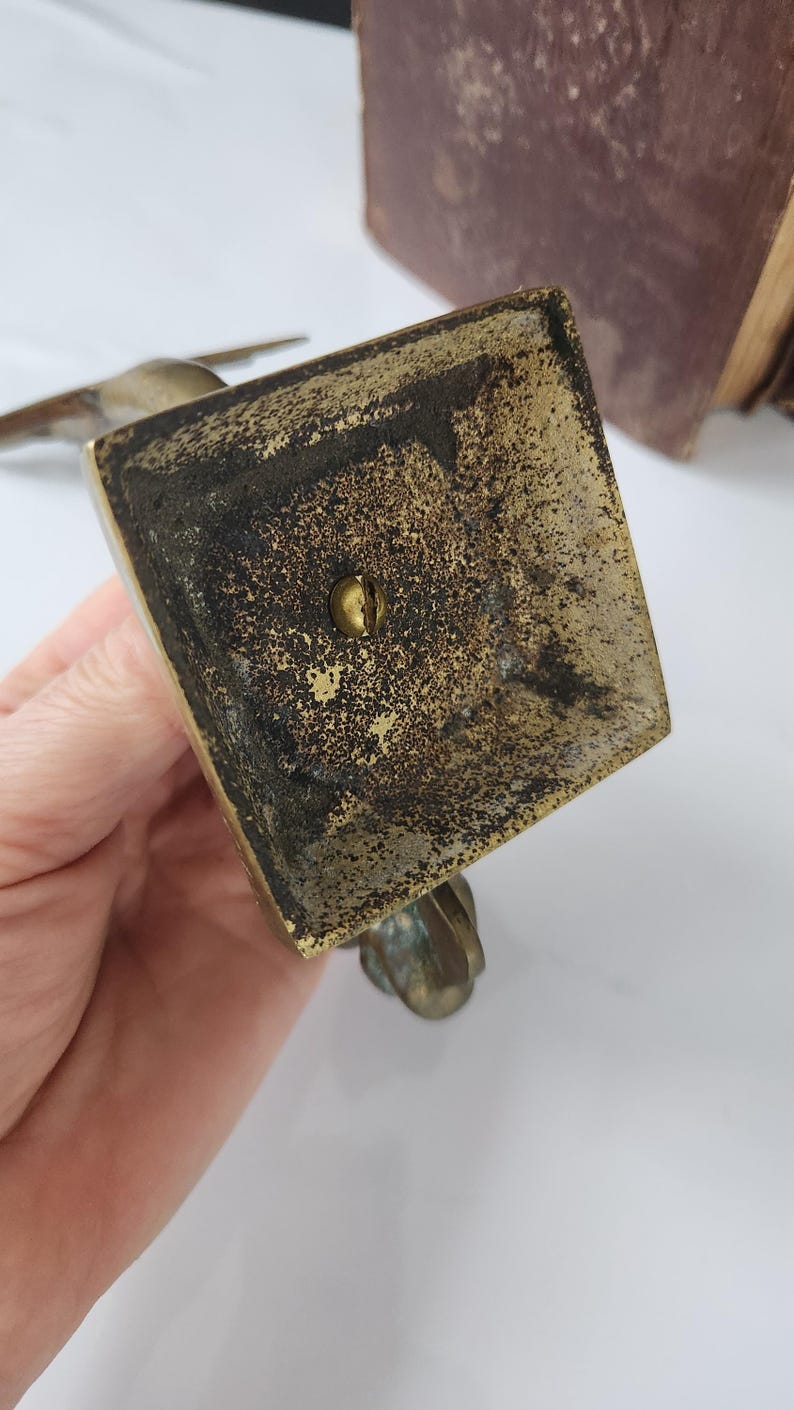 May include: A close-up view of a vintage brass bird figurine base. The square base shows signs of age with dark patina and a central screw. The bird's feet are visible, adding to the antique aesthetic. The background is blurred.
