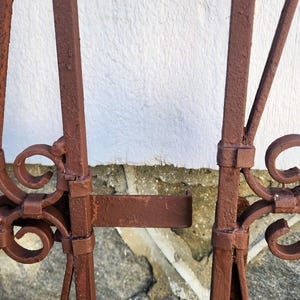 Pu&ograve; includere: Primo piano di una sezione di un cancello in metallo marrone arrugginito. Il cancello presenta barre verticali con decorazioni a volute e un chiavistello. Lo sfondo &egrave; un muro bianco strutturato e pietra.