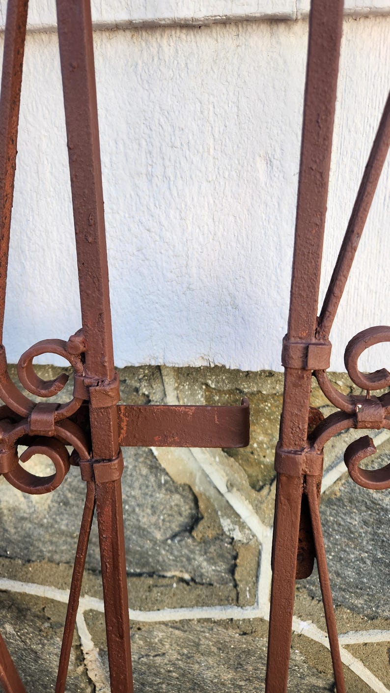 Pu&ograve; includere: Primo piano di una sezione di un cancello in metallo marrone arrugginito. Il cancello presenta barre verticali con decorazioni a volute e un chiavistello. Lo sfondo &egrave; un muro bianco strutturato e pietra.