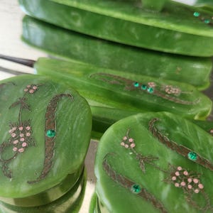 May include: A green jadeite vanity set featuring floral and leaf designs. The set includes a tray, a brush, and several oval containers. The containers are decorated with pink floral accents and green gemstone embellishments.