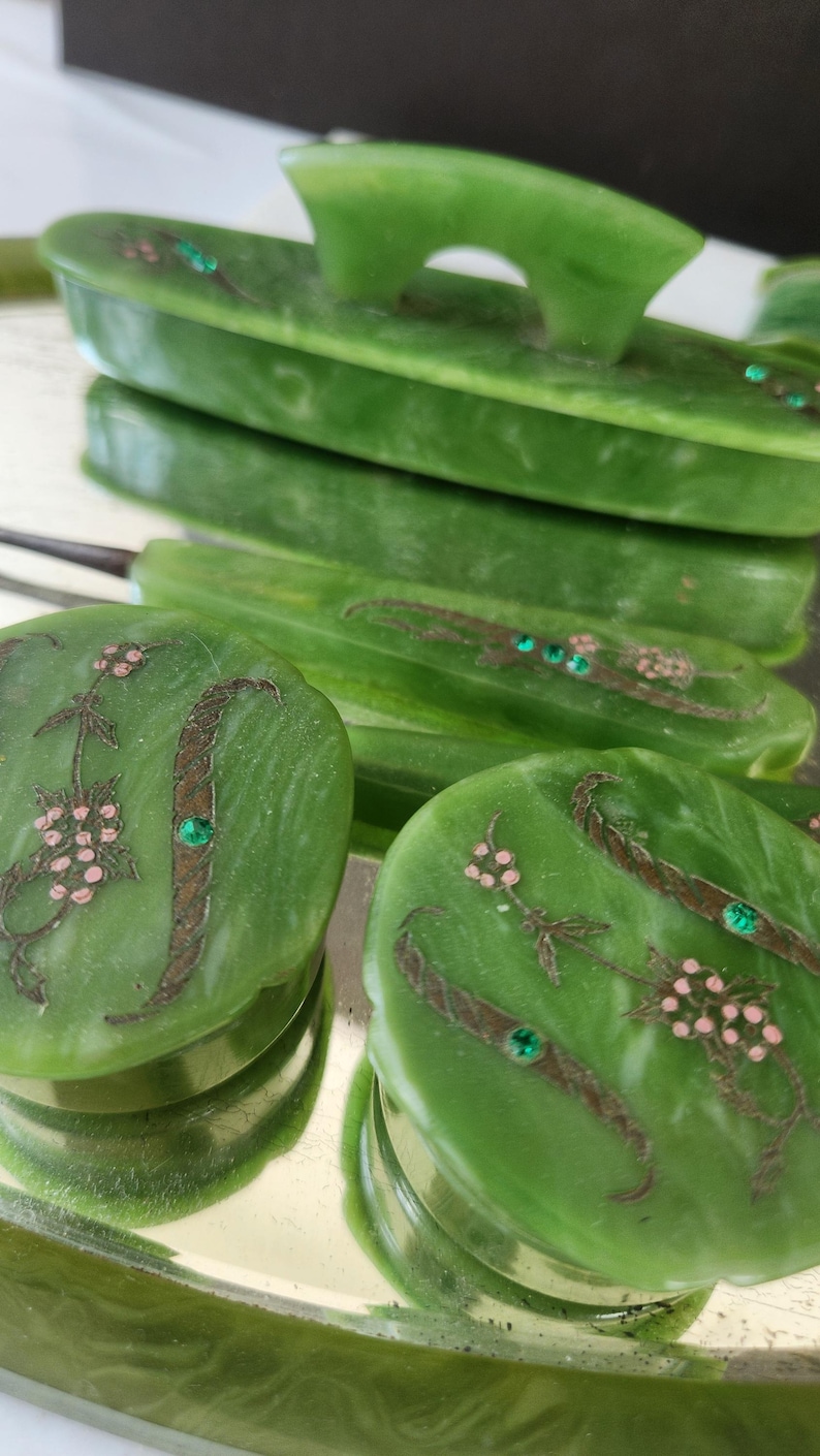 May include: A green jadeite vanity set featuring floral and leaf designs. The set includes a tray, a brush, and several oval containers. The containers are decorated with pink floral accents and green gemstone embellishments.