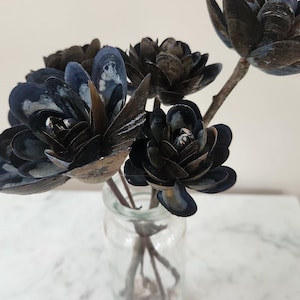 Könnte beinhalten: Ein klares Glasgefäß mit einem Strauß aus dunkelblauen und braunen Muschelblumen. Die Blumen sind in einem Bouquet angeordnet. Der Hintergrund ist eine schlichte weiße Wand.