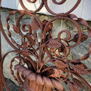 Pu&ograve; includere: Primo piano di una sezione di cancello decorativo in ferro battuto marrone arrugginito. Il design presenta elementi floreali, tra cui foglie e una fioriera centrale con fogliame. Volute e riccioli si aggiungono all'estetica ornata e vintage. Il cancello &egrave; probabilmente per uso in giardino o all'aperto.