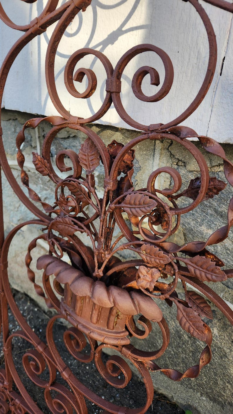 Pu&ograve; includere: Primo piano di una sezione di cancello decorativo in ferro battuto marrone arrugginito. Il design presenta elementi floreali, tra cui foglie e una fioriera centrale con fogliame. Volute e riccioli si aggiungono all'estetica ornata e vintage. Il cancello &egrave; probabilmente per uso in giardino o all'aperto.