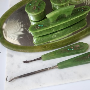 May include: A set of vintage green vanity items, including a tray, two small jars, a comb, and two manicure tools. The items are decorated with small, colourful embellishments. A green clock is also visible in the background.