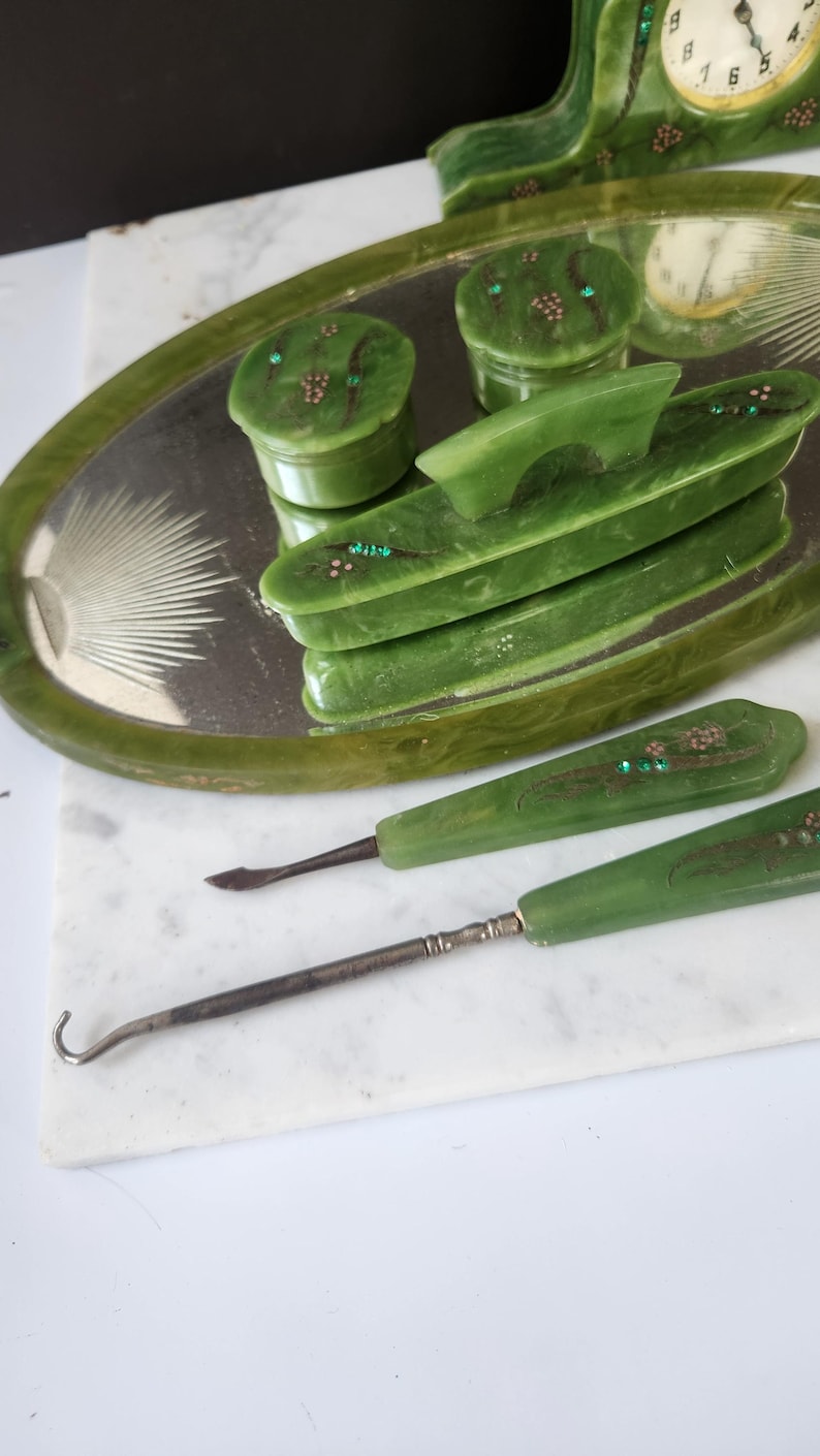 May include: A set of vintage green vanity items, including a tray, two small jars, a comb, and two manicure tools. The items are decorated with small, colourful embellishments. A green clock is also visible in the background.