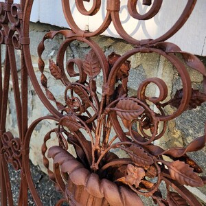 Pu&ograve; includere: Primo piano di una sezione di un cancello decorativo in ferro battuto. Il cancello presenta un'intricata lavorazione a volute, dettagli floreali e una finitura color ruggine. Un vaso con foglie &egrave; integrato nel design. Il cancello &egrave; probabilmente per giardino o esterno.