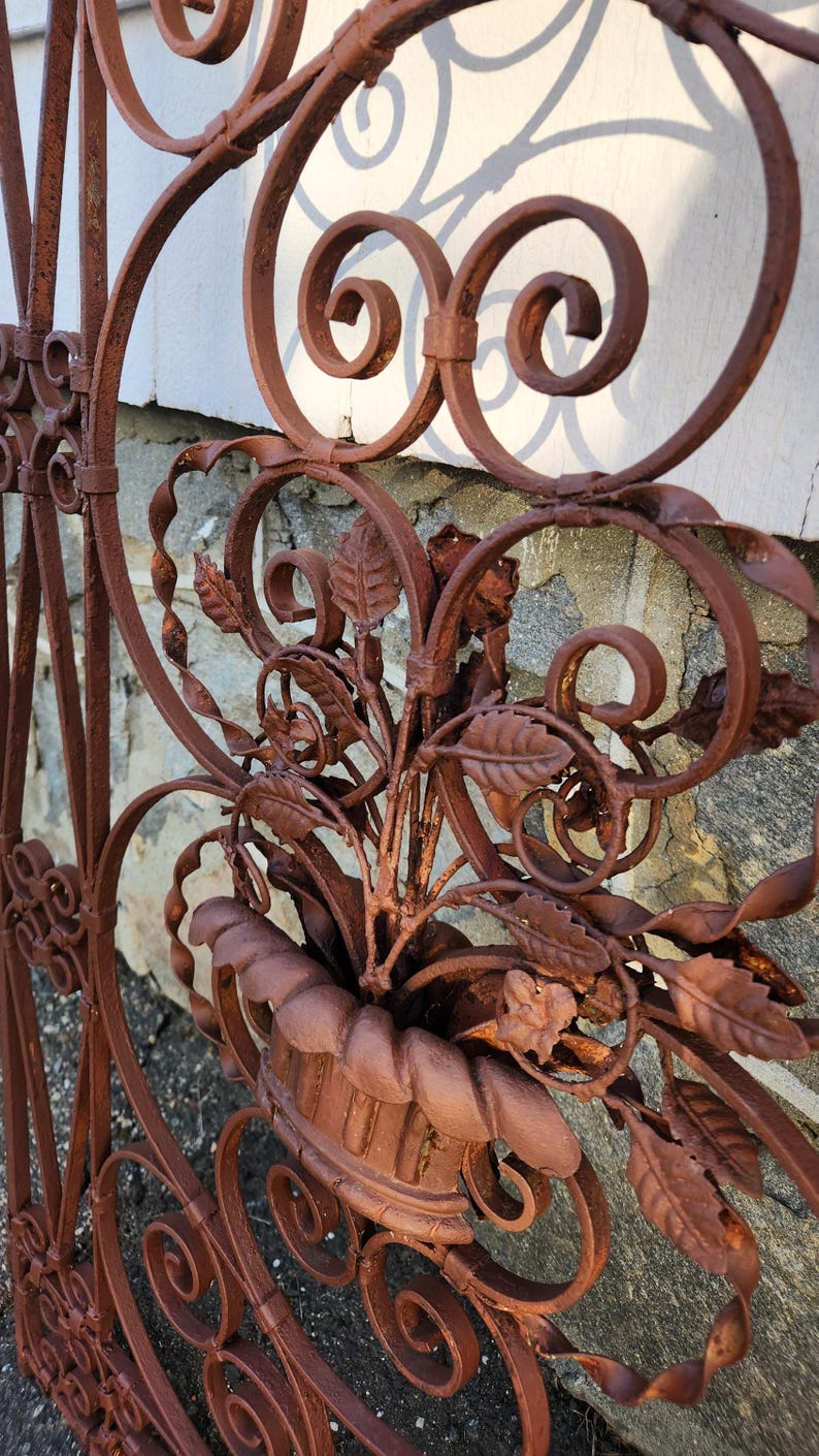 Pu&ograve; includere: Primo piano di una sezione di un cancello decorativo in ferro battuto. Il cancello presenta un'intricata lavorazione a volute, dettagli floreali e una finitura color ruggine. Un vaso con foglie &egrave; integrato nel design. Il cancello &egrave; probabilmente per giardino o esterno.