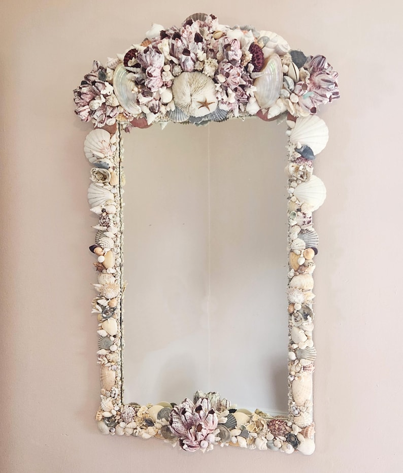 May include: A rectangular mirror with a frame intricately decorated with various seashells. The frame features a mix of white, beige, and purple shells, creating a coastal, decorative aesthetic. The top of the frame has a floral arrangement of shells.