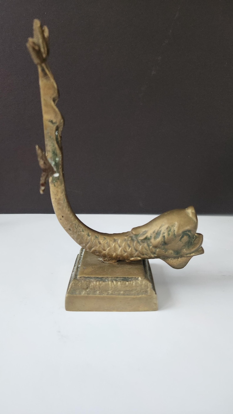 May include: A vintage brass sculpture of a fish with a curved body and a square base. The fish has detailed scales and fins, with a weathered, aged appearance. The sculpture is a decorative item.