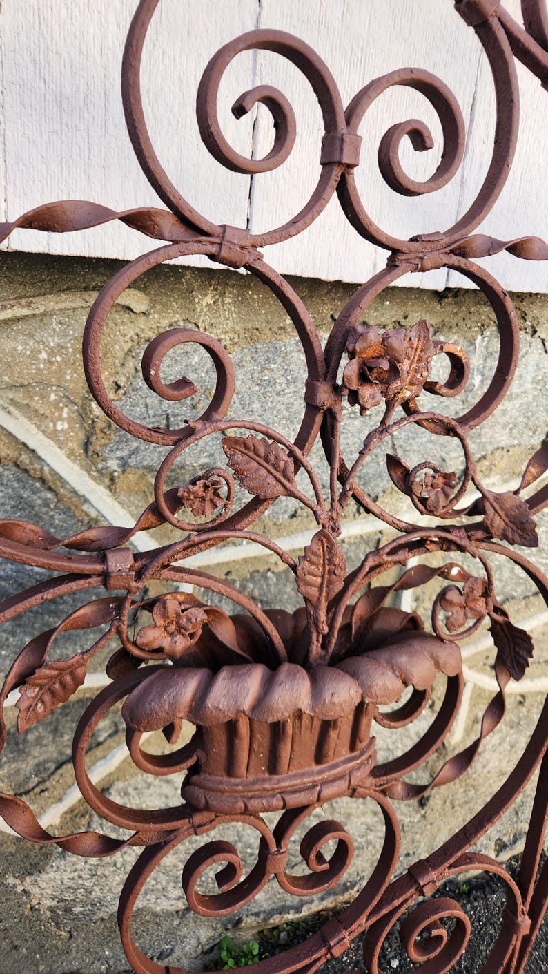 Pu&ograve; includere: Primo piano di una sezione di cancello decorativo in ferro battuto marrone arrugginito. Il design presenta elementi floreali, tra cui fiori e foglie, e accenti a spirale. Il cancello &egrave; probabilmente per uso esterno.