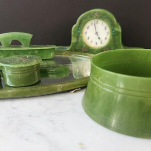 May include: A vintage green vanity set, including a clock, tray, brush, and two small jars. The set is made of a jade-like material and has a classic design. The clock has a white face with black numerals.