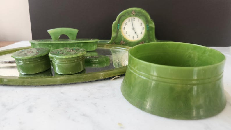 May include: A vintage green vanity set, including a clock, tray, brush, and two small jars. The set is made of a jade-like material and has a classic design. The clock has a white face with black numerals.