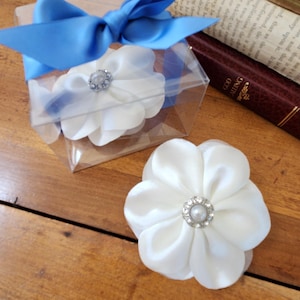 May include: White satin flower with a pearl and rhinestone center, next to a clear box with a blue ribbon. The flower is on a wooden surface, with old books in the background.