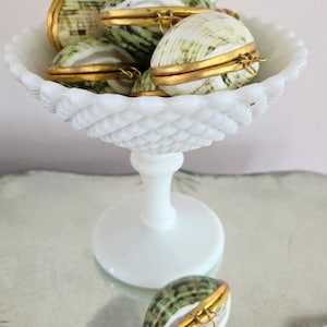 May include: A collection of small, oval-shaped shell containers with gold-toned metal hinges and rims, featuring green and white mottled patterns. Several of these hinged shell boxes are displayed inside a white milk glass pedestal bowl.
