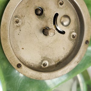 May include: Close-up of the back of a vintage clock. The clock's metal mechanism is visible, with a circular design and several screws. The clock has a green, possibly jade-like, frame. The metal components show signs of age.