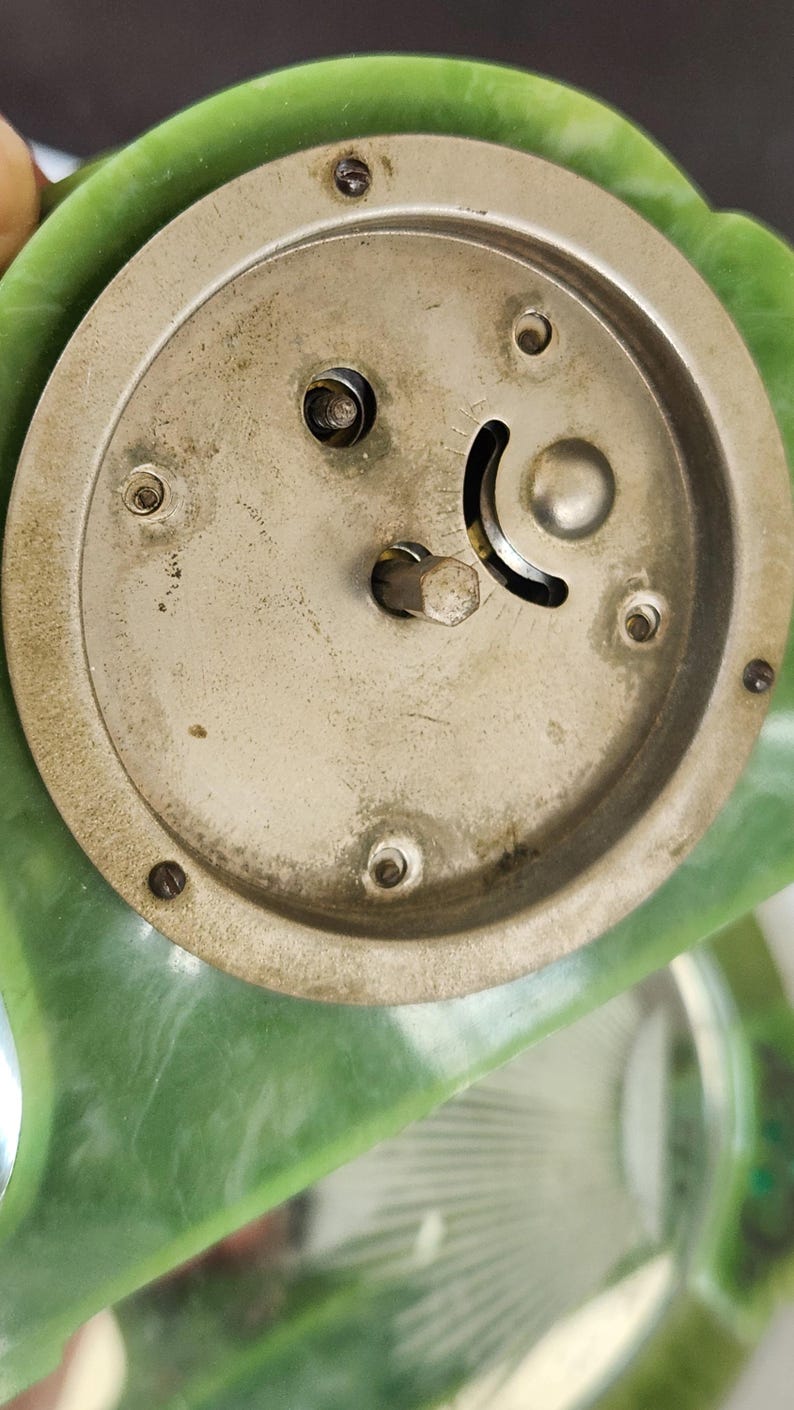 May include: Close-up of the back of a vintage clock. The clock's metal mechanism is visible, with a circular design and several screws. The clock has a green, possibly jade-like, frame. The metal components show signs of age.