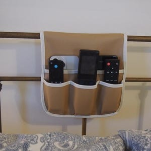 Puede incluir: Un organizador de mesita de noche beige con tres bolsillos, ribeteado en blanco, cuelga de una estructura de cama. Tres mandos a distancia negros se guardan en los bolsillos. La cama tiene almohadas estampadas.