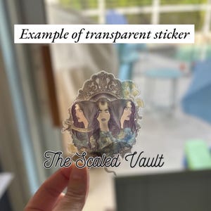 Peut inclure: Autocollant transparent repr&eacute;sentant trois figures f&eacute;minines stylis&eacute;es aux cheveux fonc&eacute;s et aux expressions intenses, encadr&eacute;es par une bordure orn&eacute;e. Le texte "Example of transparent sticker" et "The Scaled Vault" est inclus.