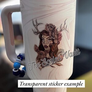 Peut inclure: Un gobelet blanc avec une poign&eacute;e et un exemple d'autocollant transparent. L'autocollant repr&eacute;sente une femme avec des bois, des fleurs et un petit animal. Le texte "The Scaled Vault" est &eacute;galement visible.