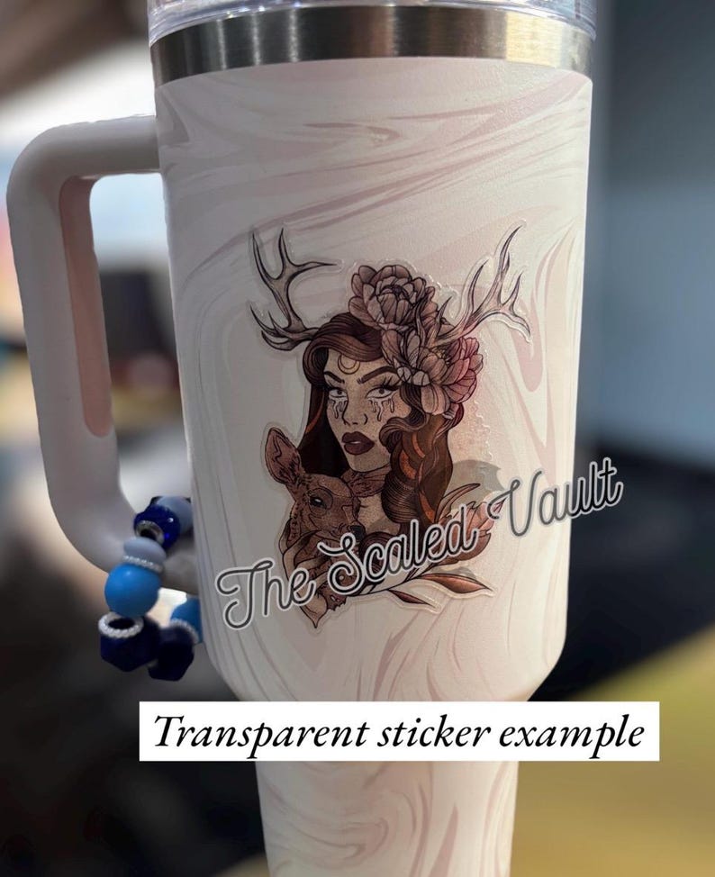 Peut inclure: Un gobelet blanc avec une poign&eacute;e et un exemple d'autocollant transparent. L'autocollant repr&eacute;sente une femme avec des bois, des fleurs et un petit animal. Le texte "The Scaled Vault" est &eacute;galement visible.