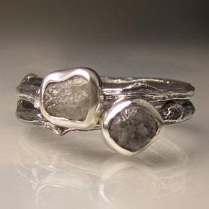 Może przedstawiać: Srebrny pierścionek z dwoma surowymi, szarymi diamentami osadzonymi w oprawie w kształcie gałązki.