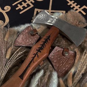 May include: A handcrafted axe with a silver blade and a wooden handle featuring a black tree design. Includes two brown leather sheaths with embossed patterns and a decorative metal emblem. The axe is a symbol of craftsmanship.