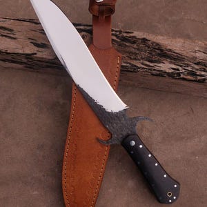 May include: A black and silver knife with a curved blade and a black handle. The knife is in a brown leather sheath with decorative stitching.