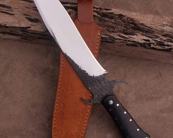 May include: A black and silver knife with a curved blade and a black handle. The knife is in a brown leather sheath with decorative stitching.