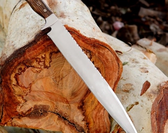 May include: A large, stainless steel knife with a serrated edge and a wooden handle. The handle is dark brown with three decorative rivets. The knife is resting on a section of a tree trunk, showcasing the wood grain.