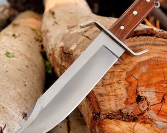 May include: A large, polished steel knife with a wooden handle and a silver guard. The knife is resting on a section of a tree trunk, showcasing its sharp blade and sturdy construction. The handle is secured with three rivets.