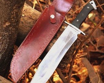 May include: A large hunting knife with a polished steel blade and a black handle. The knife is accompanied by a brown leather sheath with decorative stitching and a snap closure. The knife and sheath are displayed against a backdrop of natural wood and foliage.